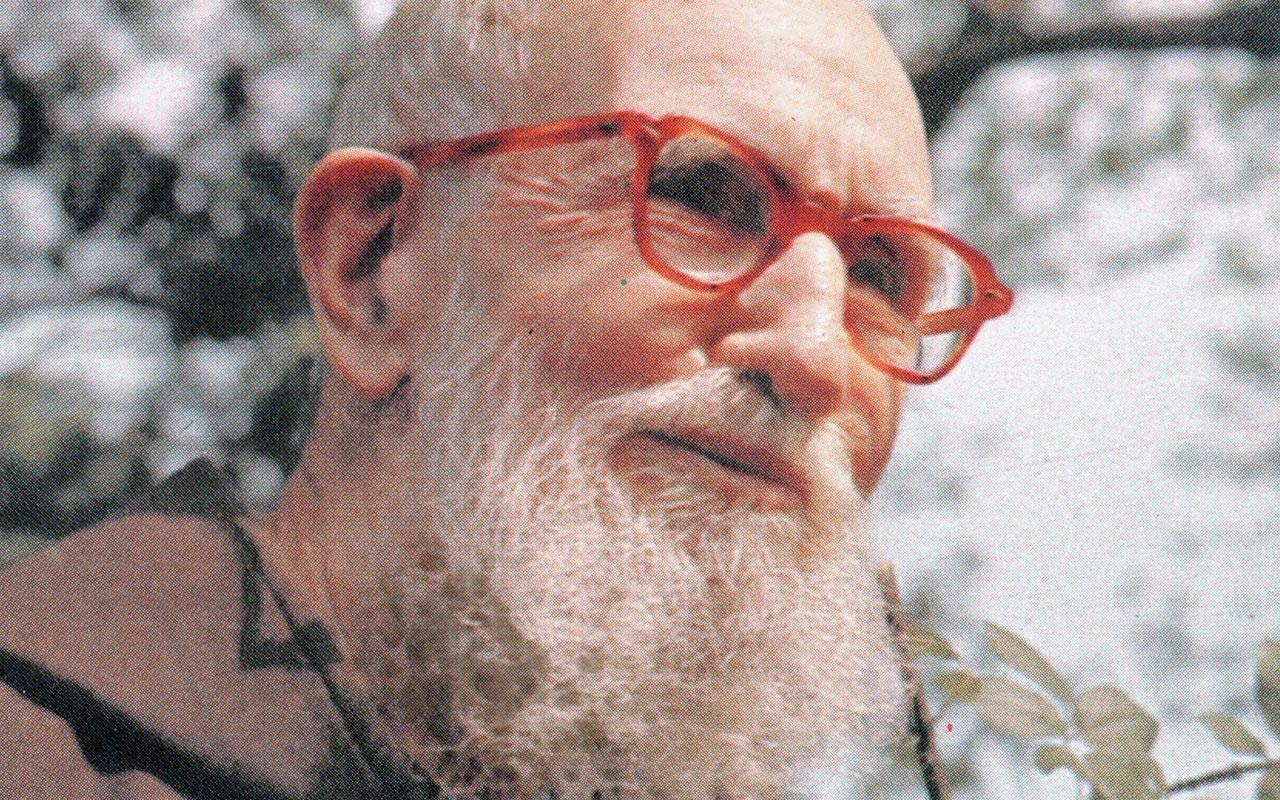 A color photograph of Blessed Solanus Casey taken outdoors. He is positioned in three-quarter profile, smiling and wearing a red set of eyeglasses.