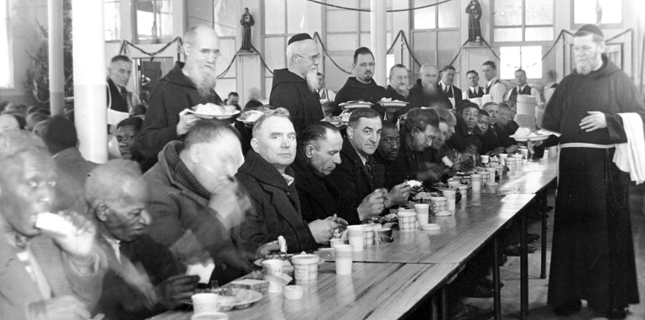 A historic photograph taken around 1940 of Blessed Solanus Casey and other Capuchin friars serving a meal at the Capuchin Soup Kitchen. Guests, all men, are seated at long tables and are eating soup and bread.