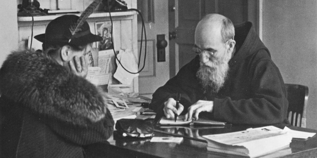A black-and-white interior photograph taken in 1941 at St. Bonaventure Monastery of Blessed Solanus Casey seated at a desk with his head down writing in a book. Seated across from him is a female visitor.