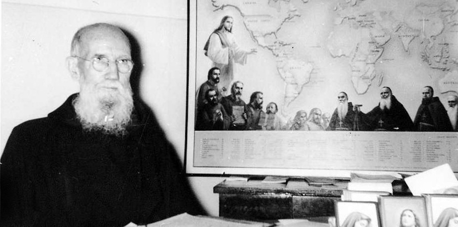 A photograph of Blessed Solanus Casey seated at a desk. Behind him on the wall is a map of Capuchin Missions around the world.