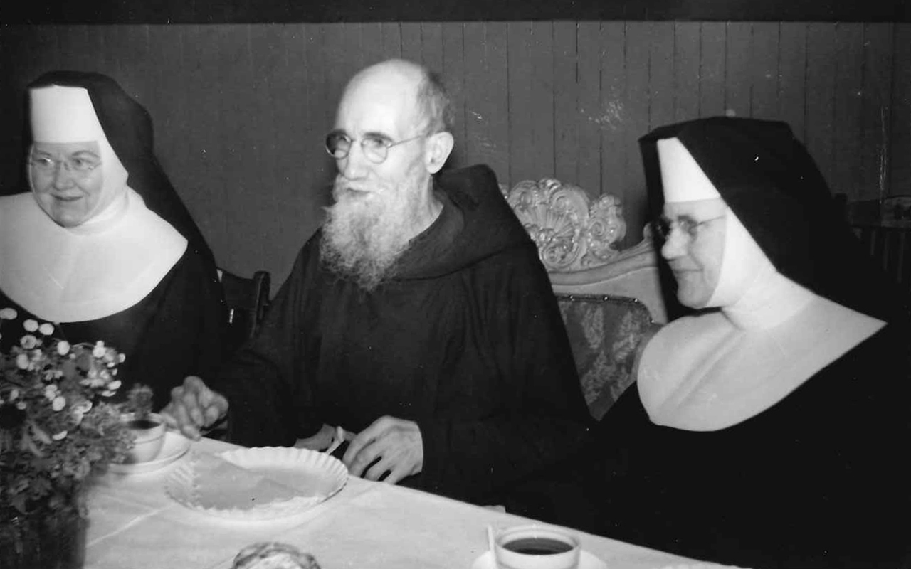 A historic photograph of Blessed Solanus Casey at a dining table flanked on each side by religious sisters wearing habits.