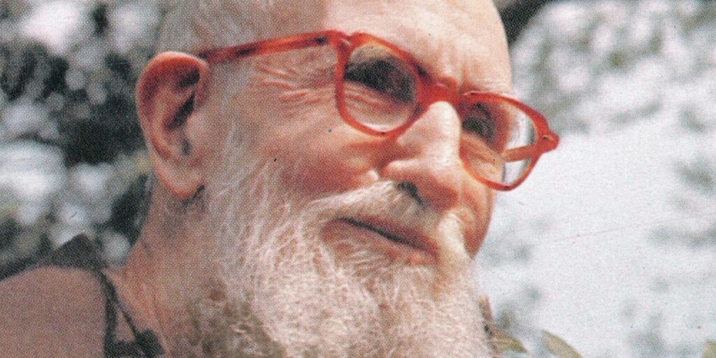 A close-up color photograph of Blessed Solanus Casey shot outdoors. He is in a three-quarter profile, wearing a pair of red eyeglasses and smiling.
