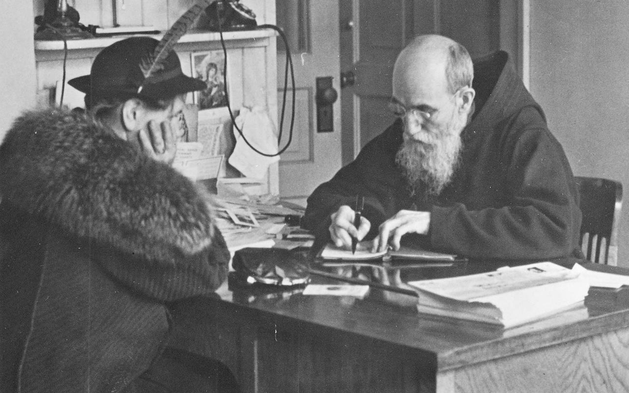 A black-and-white interior photograph taken in 1941 at St. Bonaventure Monastery of Blessed Solanus Casey seated at a desk with his head down writing in a book. Seated across from him is a female visitor.