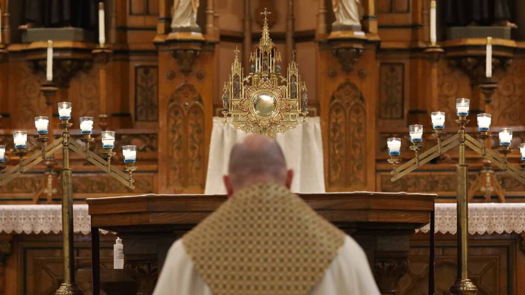 Photograph of a Capuchin Franciscan friar kneeling in front of the altar. On the altar is the Most Blessed Sacrament within a gold monstrance. Candelabra are lit and placed on each side of the monstrance.