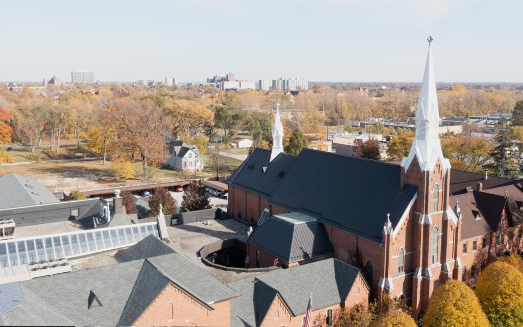 An aerial exterior photograph taken October 2022 of the Solanus Casey Center and St. Bonaventure Chapel and Monastery in the foreground. In the midground is the Islandview neighobrhood and in the background are high-rise condominiums in Detroit's Gold Coast neighbrhood.