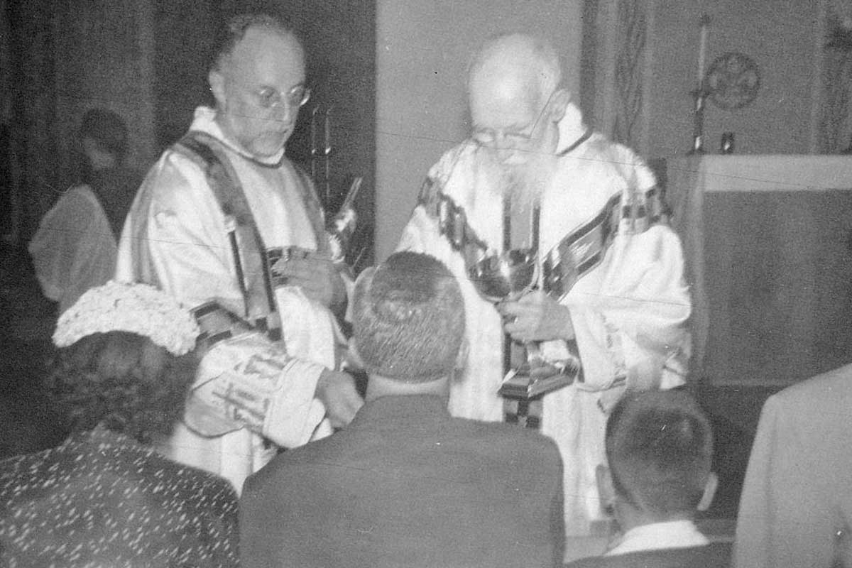 A black-and-white historic photograph of Blessed Solanus Casey distributing the Eucharist at Mass.