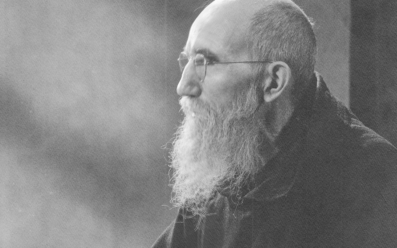 A historic black-and-white studio portrait photograph of Blessed Solanus Casey taken circa 1945 in Seattle. Blessed Solanus is wearing his habit and a pair of spectacles and looks to the left. He is holding an open prayer book in his hands.