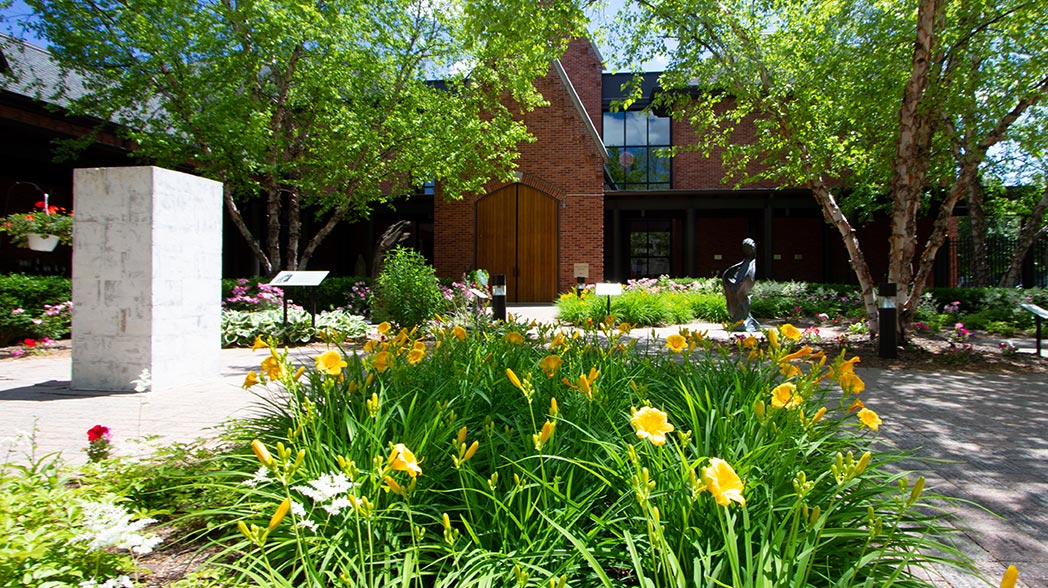 Summer photograph of the exterior of the Solanus Casey Center and the creation garden. Day lillies are in full bloom in the foreground. The garden appears lush and verdant.